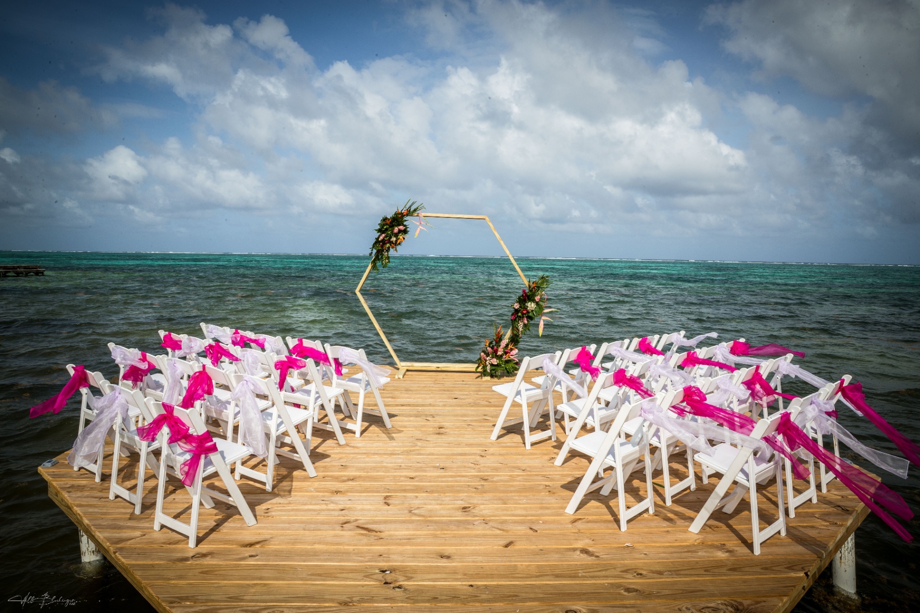 Hexagonal floral arch and white chairs with pink sashes set up on the dock over turquoise water