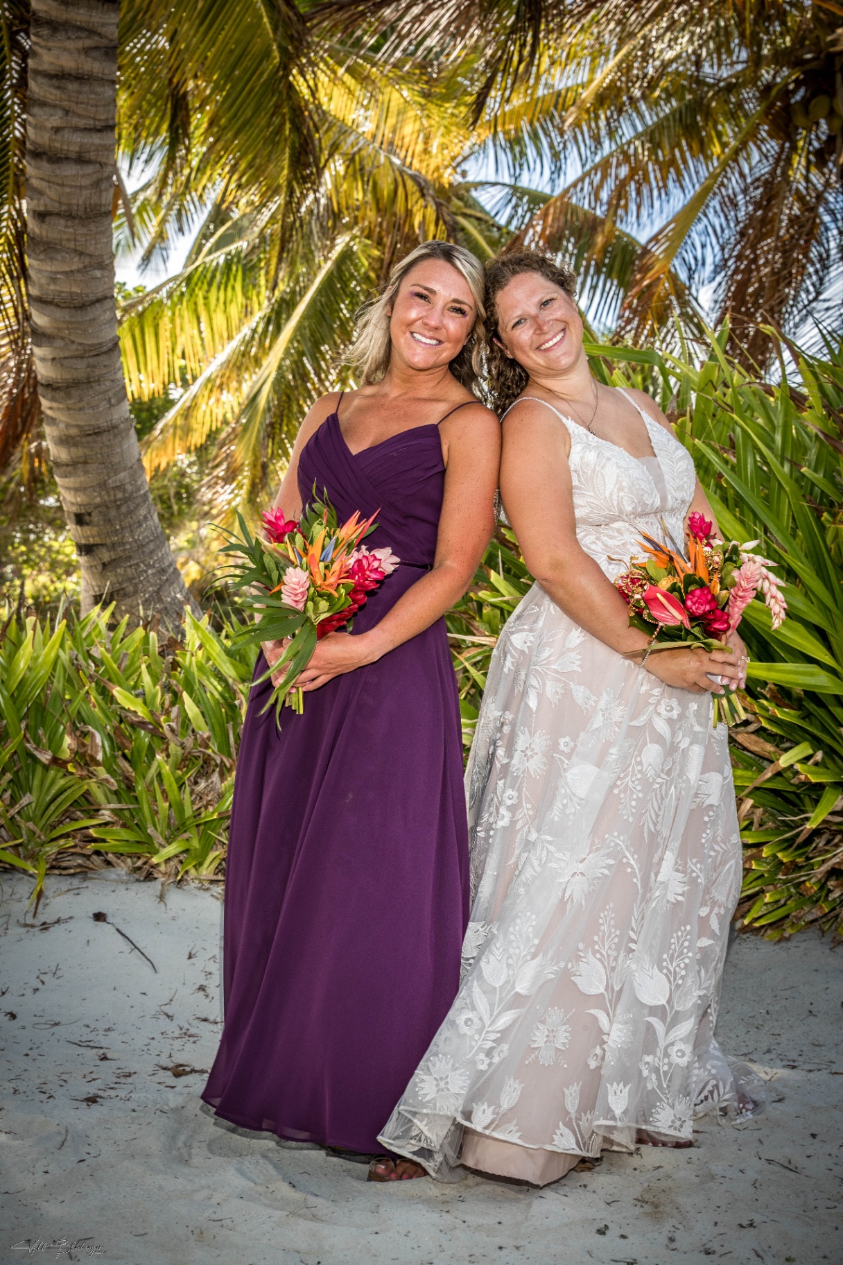 Bride and maid of honor posing back to back with their tropical bouquets under the palms